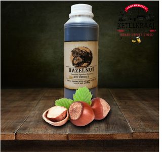 A bottle of hazelnut nut extract with a label depicting a hazelnut is placed on a wooden surface. In front of the bottle, three hazelnuts, one of which is split open to reveal the kernel, rest on the surface alongside green leaves. A dark, textured background with the logo "KETELKRAAL - BRAAI-SUPPLY STORE" is visible.