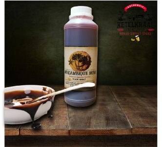 A plastic bottle labeled "Mozambique Rum Premium Signature Essence" is placed on a wooden surface beside an ashtray containing a dark liquid with a spoon. A dark backdrop with a logo reading "Authentic Ketelkraal Distillery Braai-Supply Store" enhances the rustic setting.