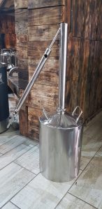 A stainless steel distillation apparatus with a tall column and angled condenser tube, standing on a tiled floor against a rustic wooden wall background.