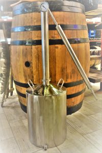 A stainless steel distillation apparatus with a thermometer and condenser tube, positioned in front of a traditional wooden barrel.